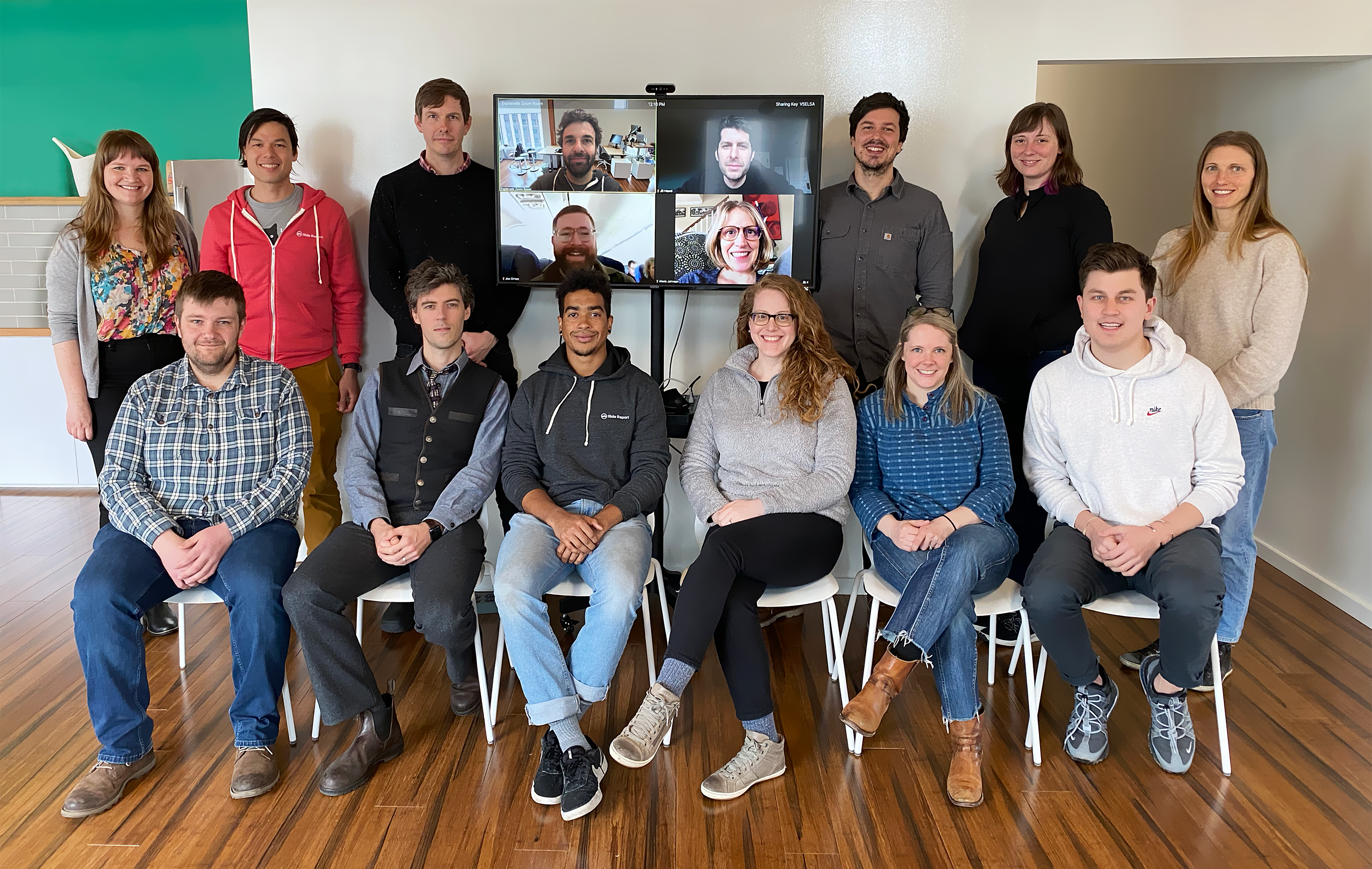 12 people in 2 rows, the first row sitting on chairs, the second row standing around a flat television screen where 4 people are pictured who have video conferenced in