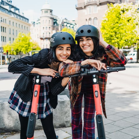 Two women on e-scooters, wearing helmets